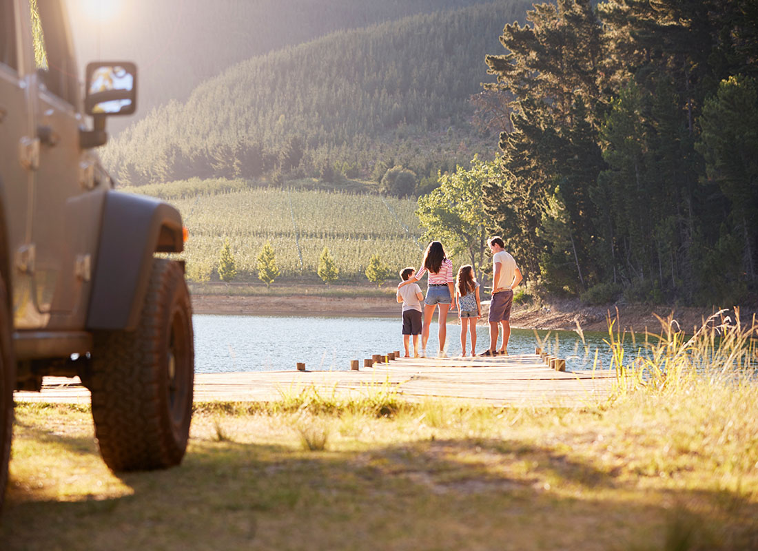 Insurance Solutions - Rear View of a Family with Two Children Standing on a Wooden Dock by the Lake with Their Jeep Parked on the Grass During a Summer Road Trip