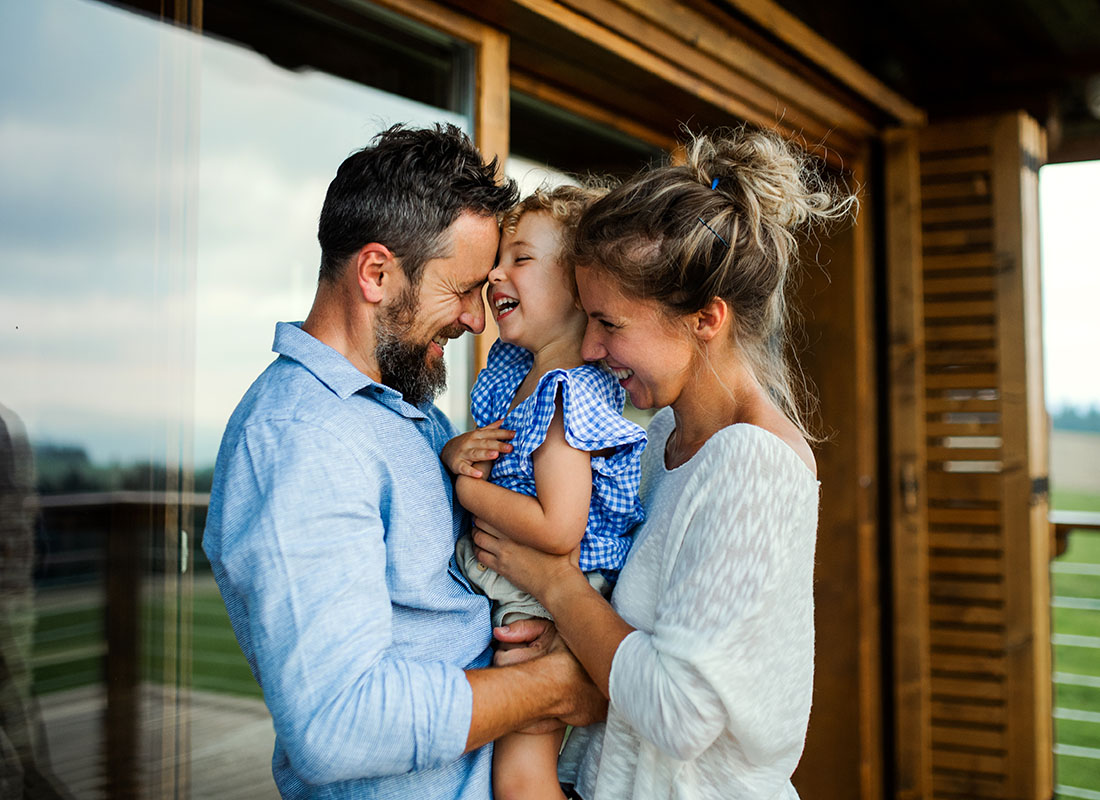 Personal Insurance - Closeup View of Cheerful Parents Cuddling and Holding Their Excited Young Daughter While Standing Outside a Cabin Style Home
